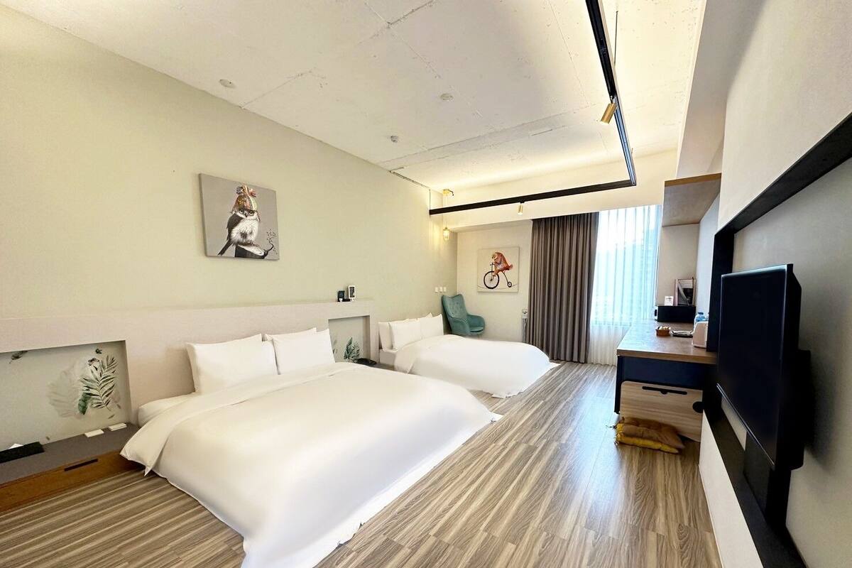Quarto de hotel com duas camas, uma escrivaninha, televisão e obras de arte nas paredes.