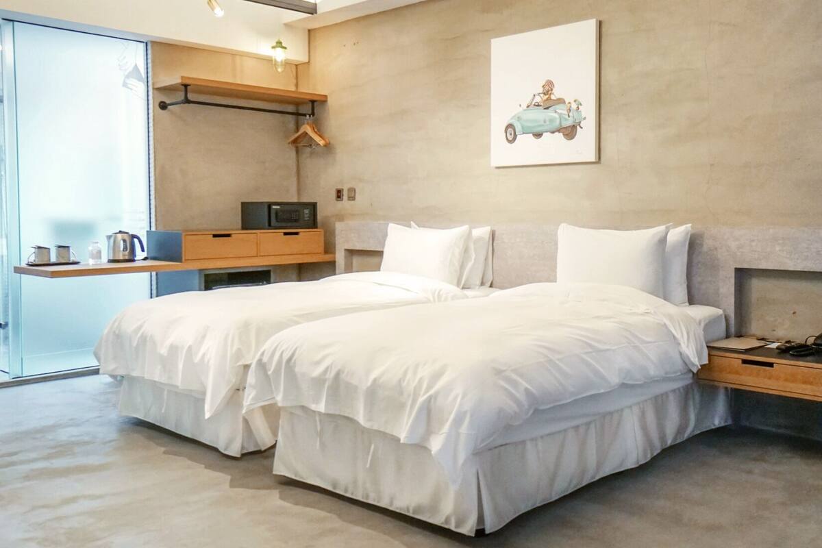 Quarto de hotel moderno com duas camas, uma mesinha de cabeceira de madeira, um micro-ondas e um cabideiro com roupas.