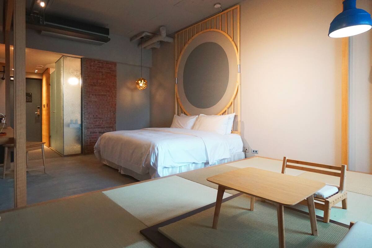 Quarto moderno com cama, mesa de madeira e uma cadeira. Há um espelho grande na parede e uma luminária pendente azul.