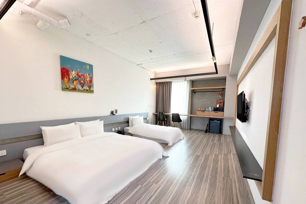 Quarto de hotel com duas camas, uma escrivaninha, uma cadeira, uma televisão e um quadro na parede.