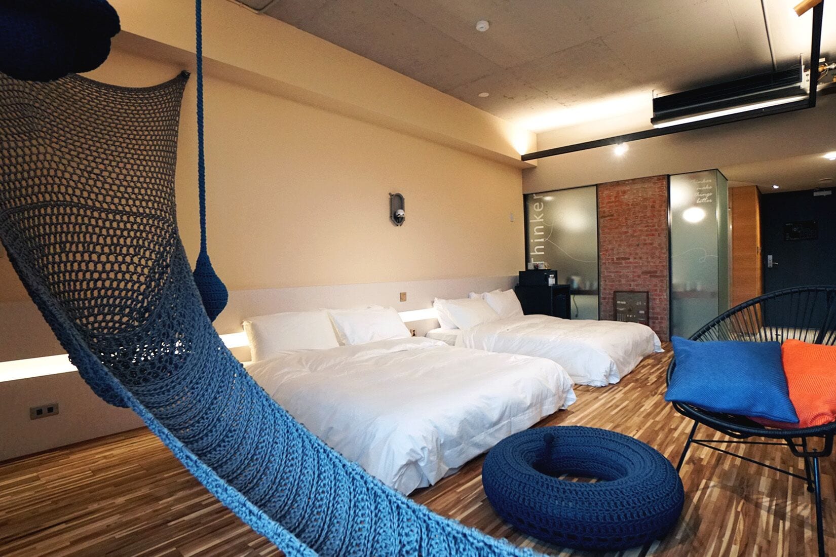 Hotellihuone, jossa on kaksi sänkyä, sininen riippumatto, sininen ja oranssi tuoli sekä tiiliseinä.