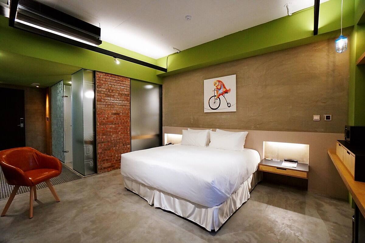 Quarto de hotel moderno com uma cama grande, uma cadeira vermelha, uma mesa com abajur e um quadro de bicicleta na parede.