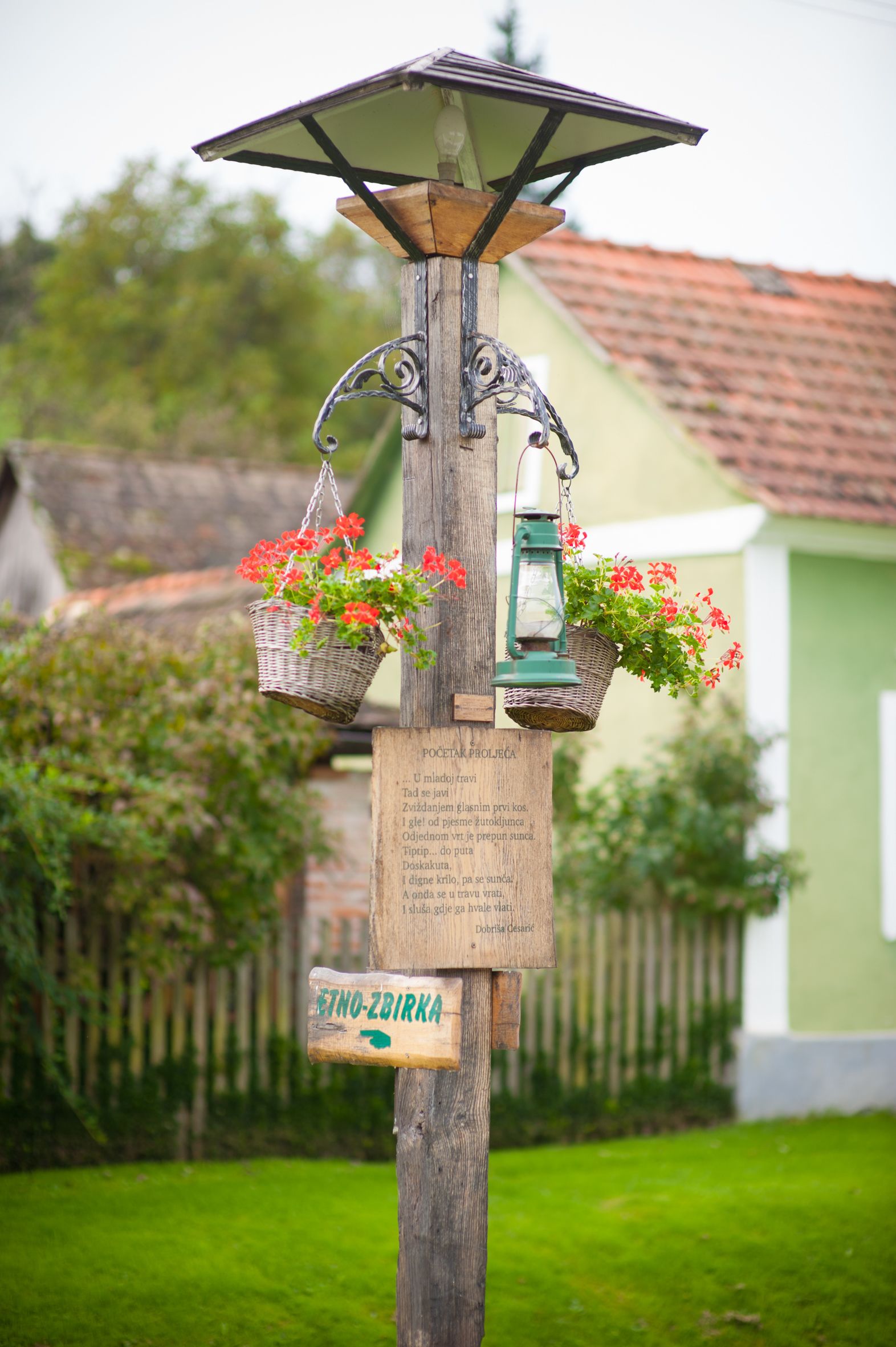 Ein hölzernes Wegweiser mit einer Solarzelle, zwei Ampelförmchen mit roten Blüten und einer grünen Laterne.