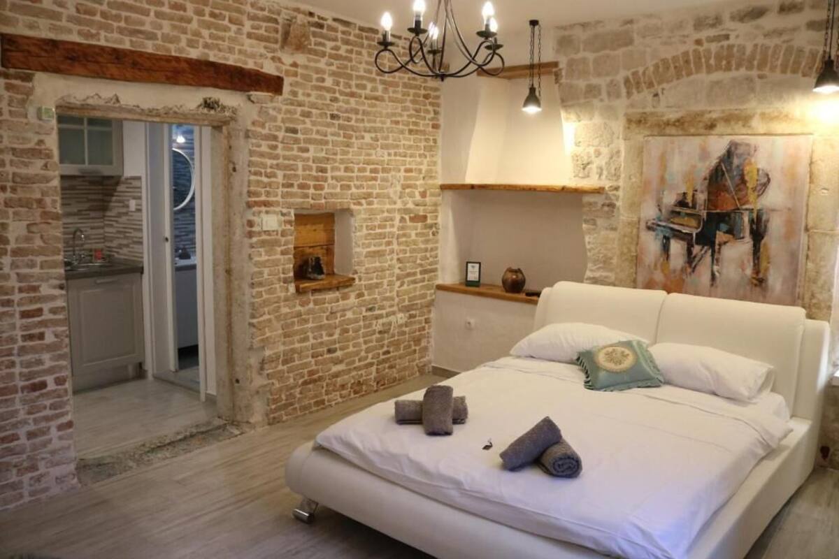 Une chambre avec un mur en briques, un lit blanc, un lustre et un tableau accroché au mur.