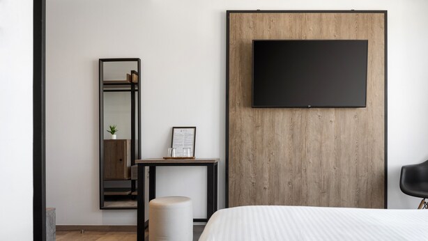A modern hotel room with a large bed, a mounted TV, a wooden accent wall, a small table with a plant, and a mirror. - Sette Suites & Rooms - Adults Only (Xylokastro-Evrostina)
