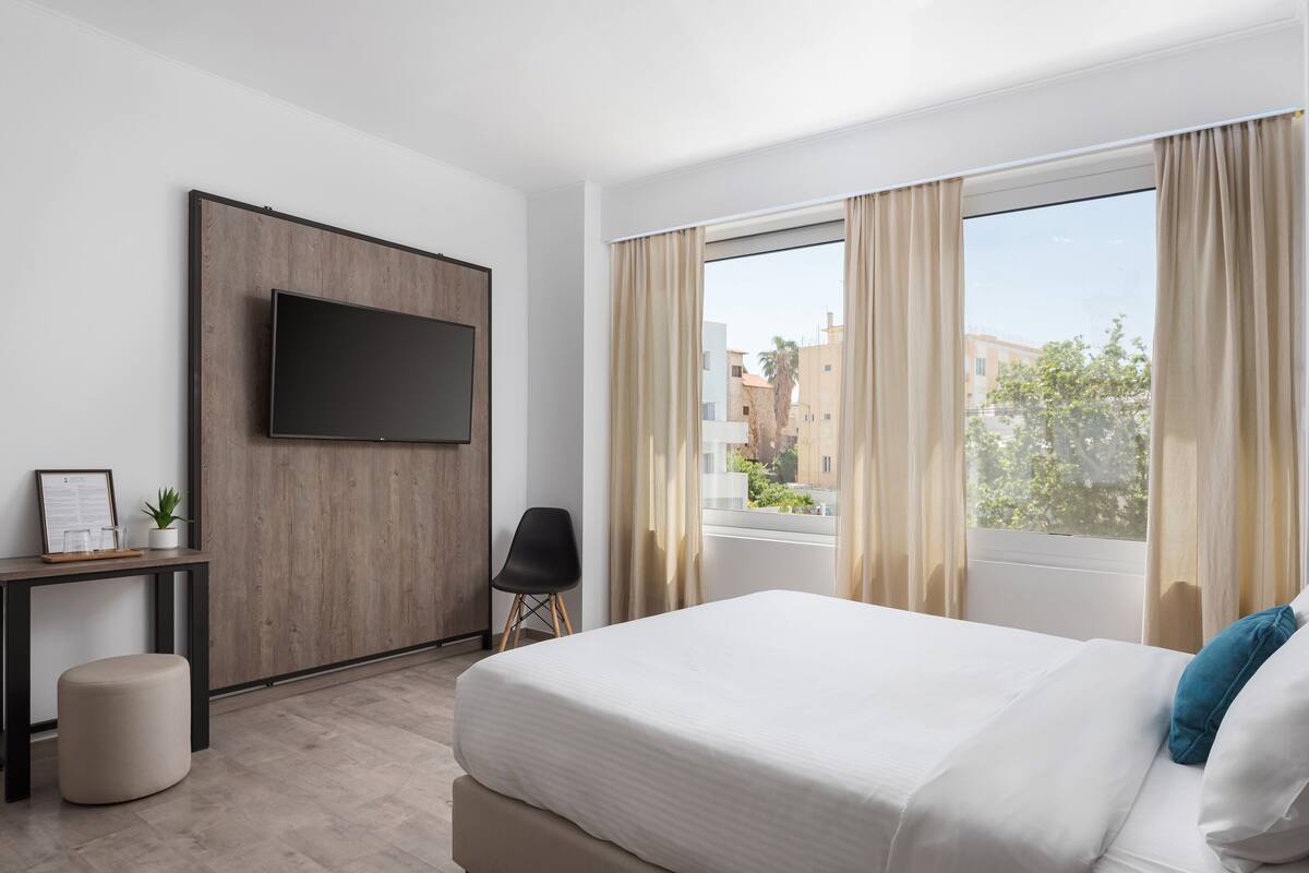 A hotel room with a bed, a TV mounted on the wall, a desk with a chair, and a window with curtains.