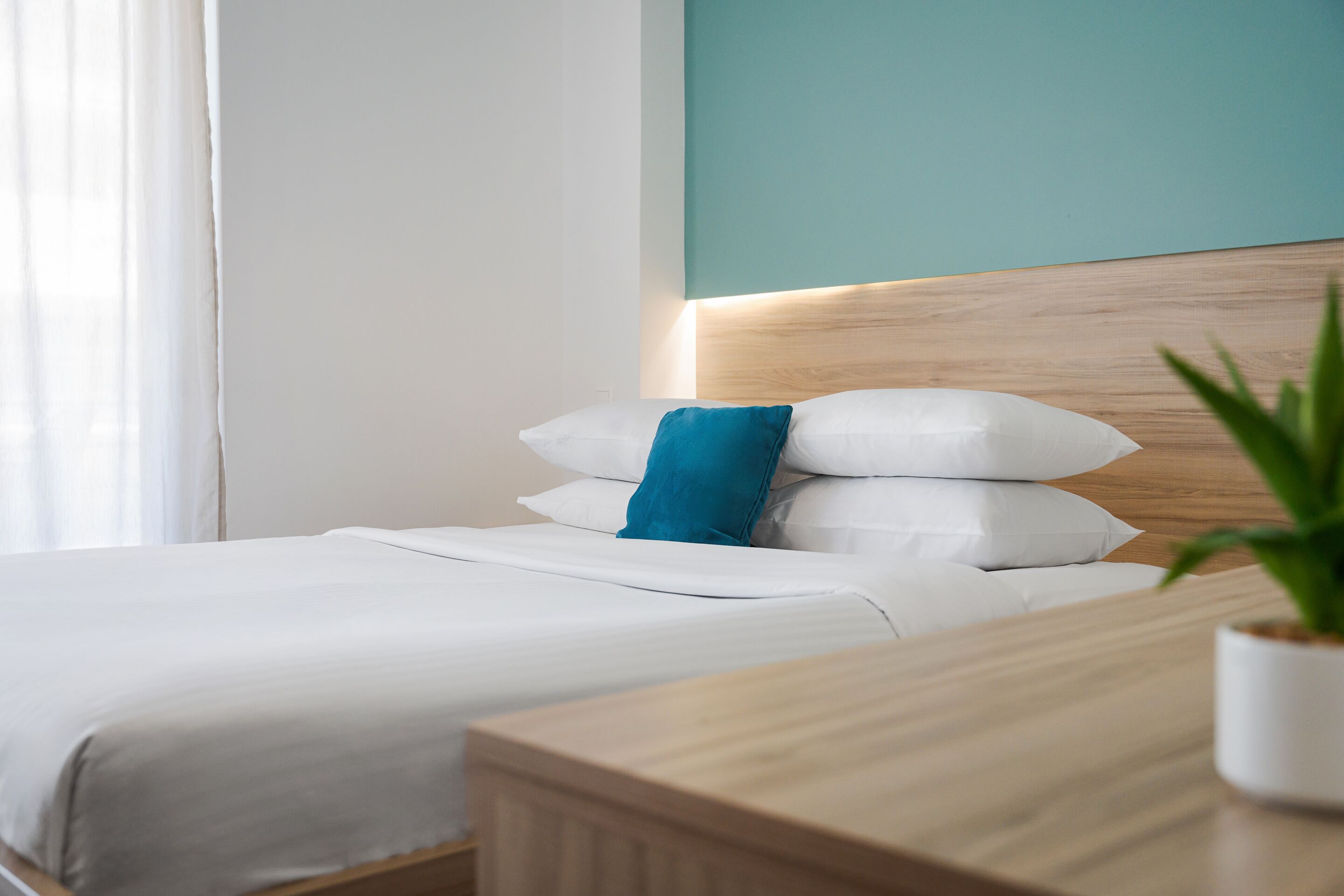 Una cama bien hecha con almohadas blancas y azules, un cabecero de madera y una mesita de noche con una planta en maceta.