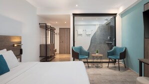 Une chambre d’hôtel moderne avec un grand lit, deux fauteuils, une petite table, un espace douche et une armoire en bois.