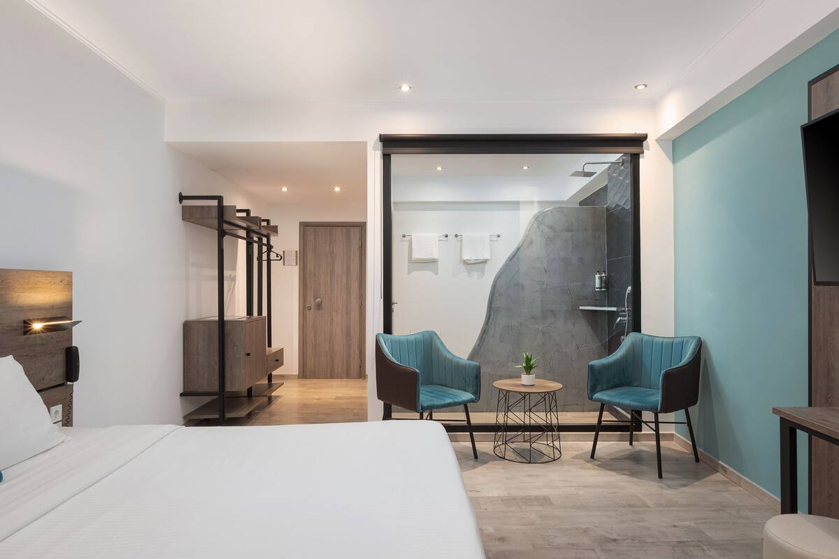 A modern hotel room with a large bed, two chairs, a small table, a shower area, and a wooden wardrobe.