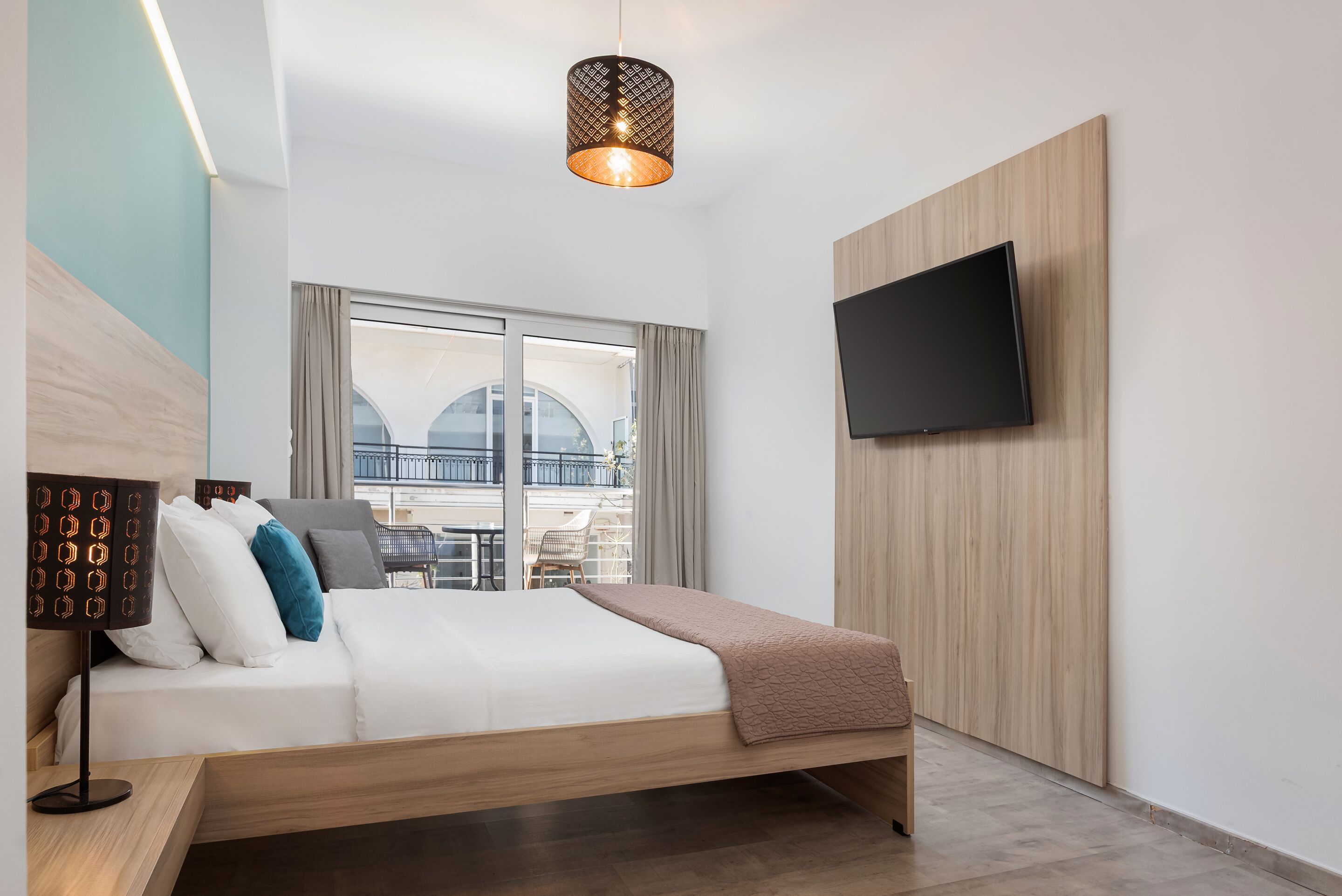 A modern hotel room with a bed, a flat-screen TV mounted on the wall, a large window with a view of the city, and a unique pendant light.