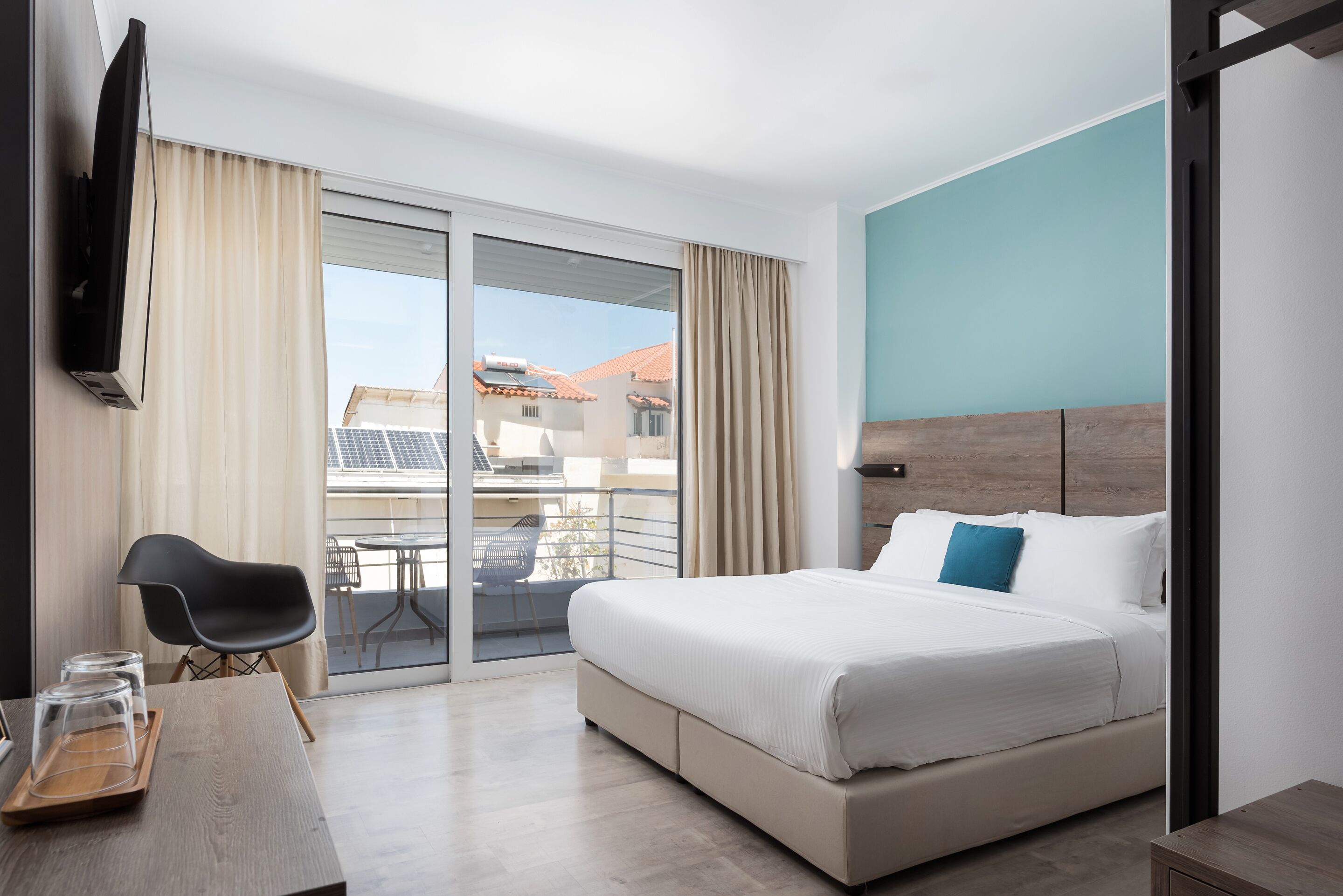 A modern hotel room with a large bed, a flat-screen TV, a dining table with chairs, and a balcony with a view of buildings and solar panels.