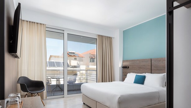 A modern hotel room with a large bed, a flat-screen TV, a dining table with chairs, and a balcony with a view of buildings and solar panels. - Sette Suites & Rooms - Adults Only (Xylokastro-Evrostina)