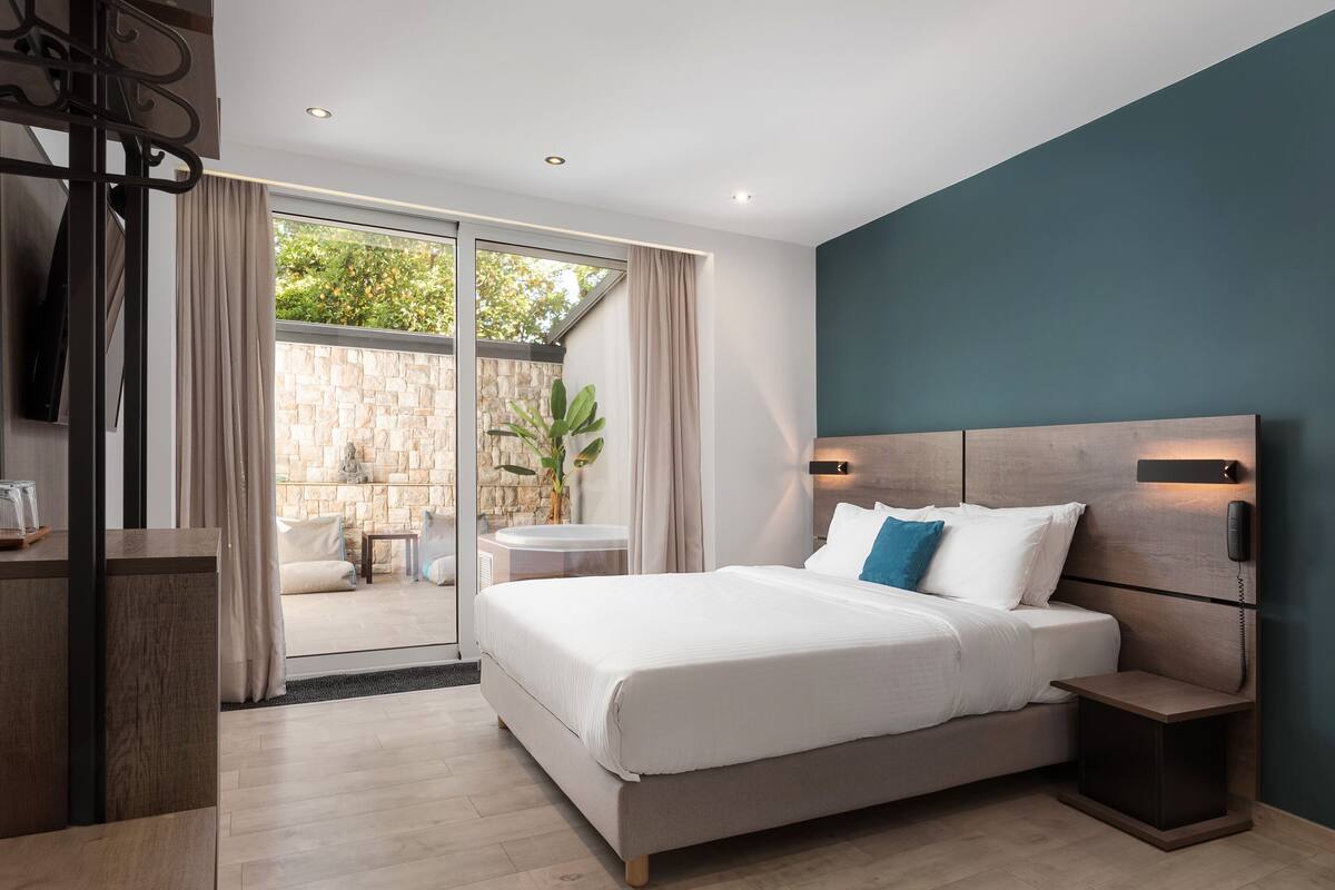 A modern hotel room with a large bed, a small bedside table, a built-in TV, and a view of the outdoor area.