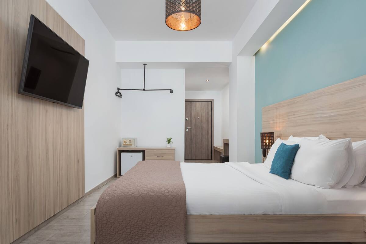 A modern hotel room with a large bed, a flat-screen TV mounted on the wall, a wooden headboard, and a small bedside table with a lamp.