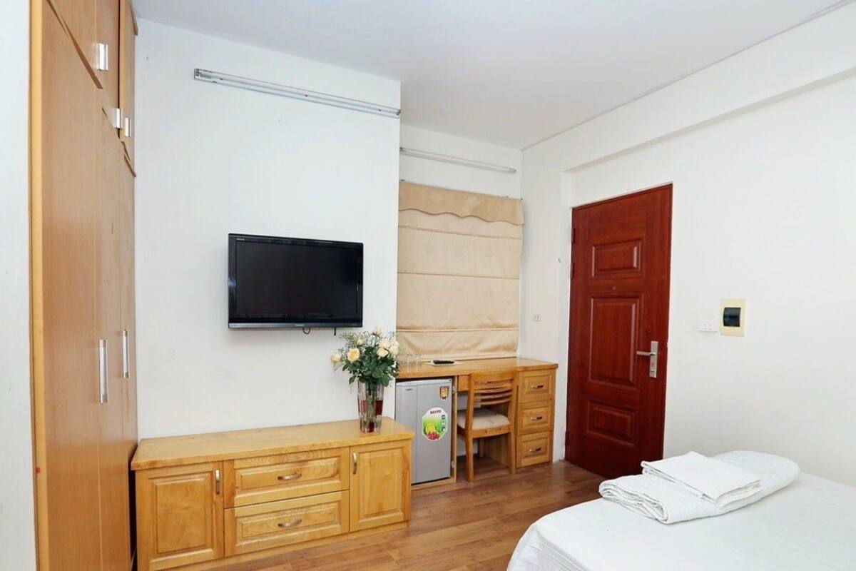 A hotel room with a bed, a desk with a chair, a television mounted on the wall, a wardrobe, and a door.