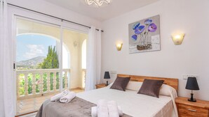 A bedroom with a large bed, a balcony with a view of greenery and a mountain, two bedside lamps, and a painting on the wall. - Lucy Moraira House for 6 people (Moraira)