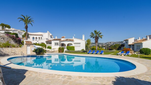 A swimming pool surrounded by stone tiles, with blue lounge chairs and palm trees. - Lucy Moraira House for 6 people (Moraira)