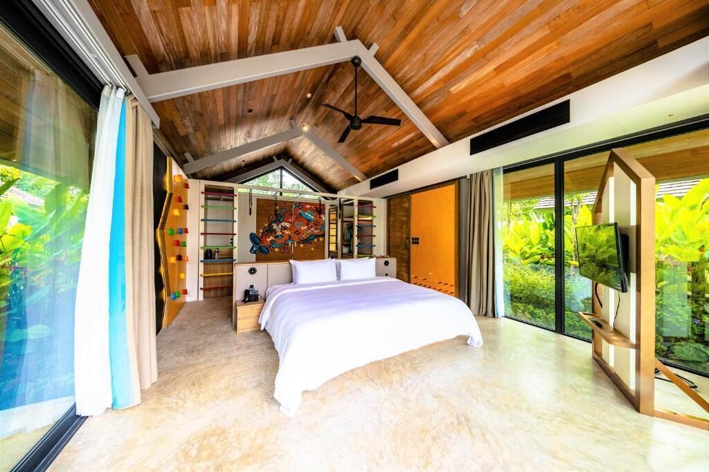 A bedroom with a large bed, wooden ceiling, and a view of greenery through large windows.