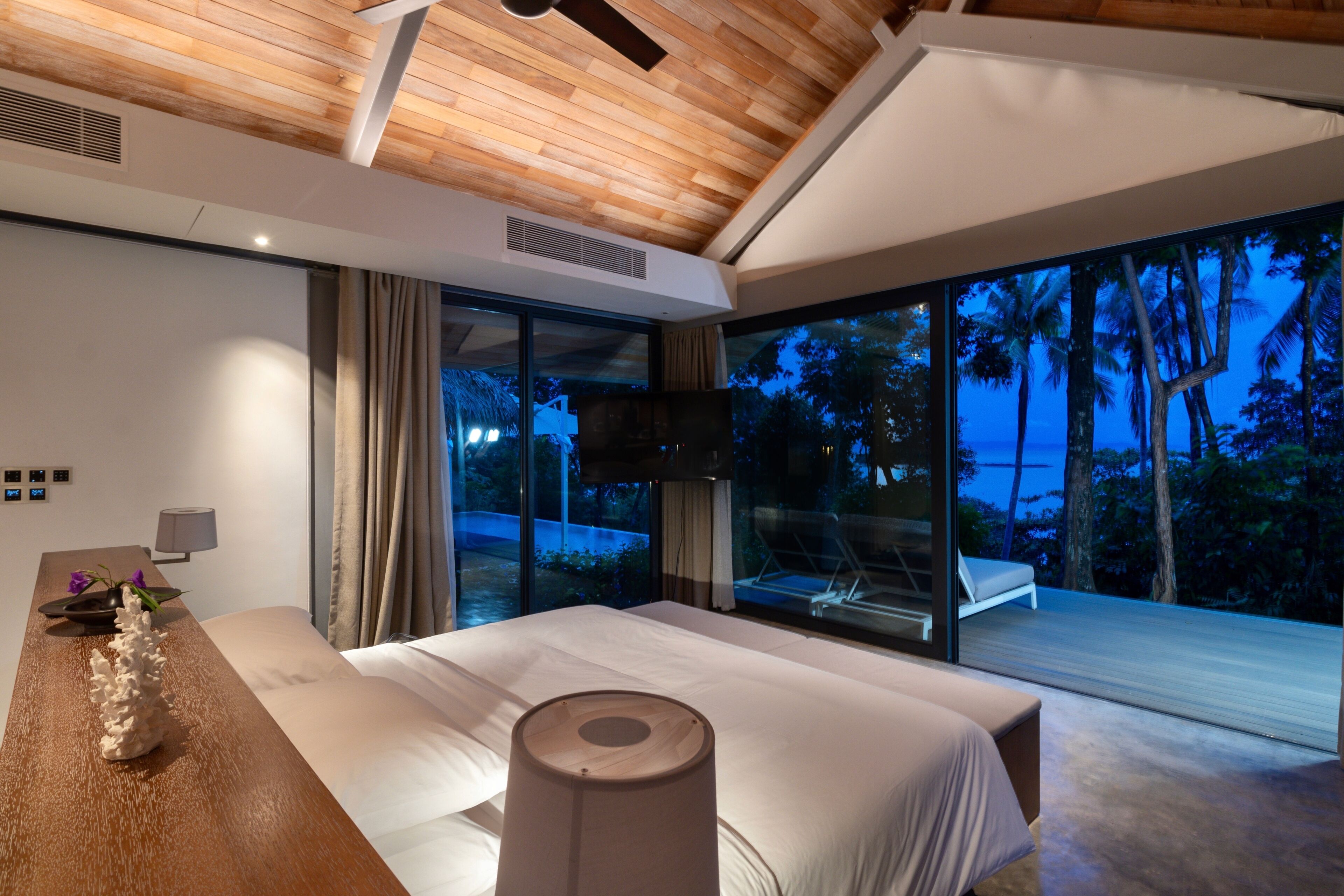 Een moderne slaapkamer met een groot bed, een houten plafond en uitzicht op de oceaan in de schemering.