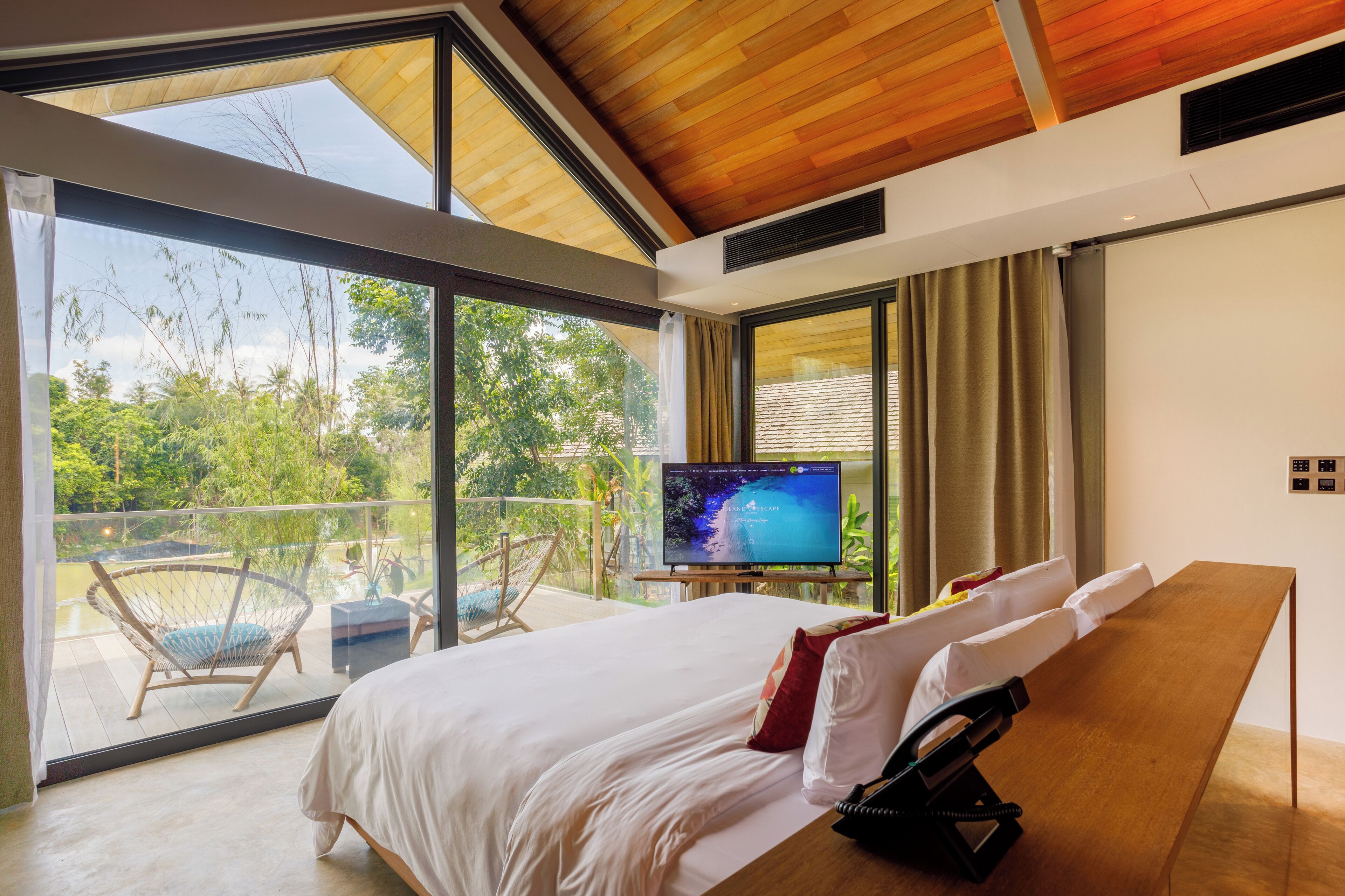 A modern hotel room with a large bed, a wooden desk, a television, and a balcony with outdoor seating.