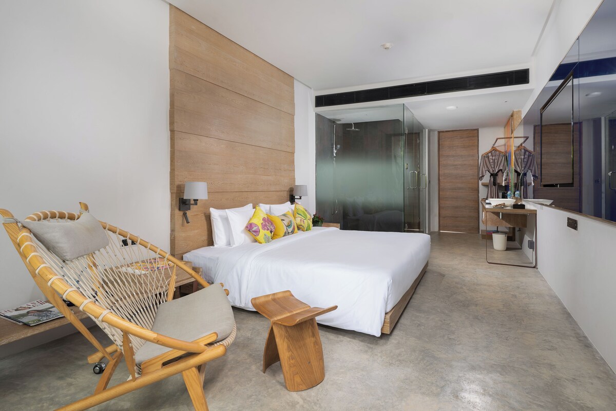 Una habitación de hotel moderna con una cama grande, una silla de madera, una mesita de madera, un espejo y una cabina de ducha de vidrio.