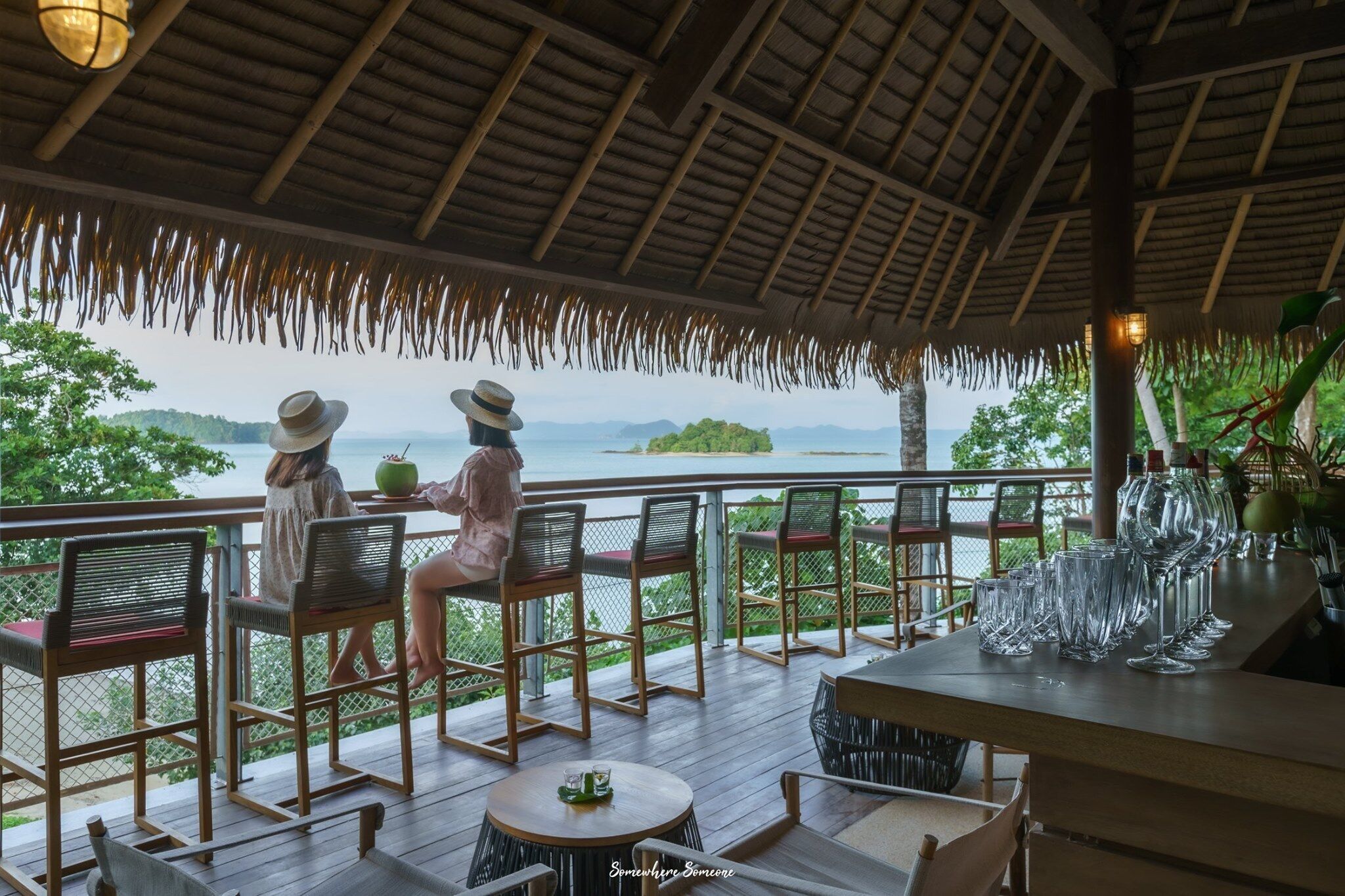 A covered outdoor seating area with wooden chairs and tables, overlooking a body of water and greenery.