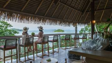 A covered outdoor seating area with wooden chairs and tables, overlooking a body of water and greenery.