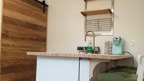 Eine kleine Küche mit einer Holztür, die sich zum Verschieben lässt, einer weißen Arbeitsplatte, einem Spülbecken und einem Stuhl mit grüner Sitzauflage.