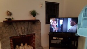 A living room with a stone fireplace, a television on a stand, and a window with blinds. - Cozy stand alone cottage with Bermuda Character  (St.George's, Bermuda)