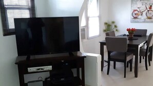 A living room with a flat-screen TV on a dark wooden stand, a dining area with a table and chairs, and a poster on the wall. - Cozy stand alone cottage with Bermuda Character  (St.George's, Bermuda)
