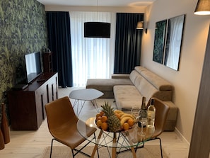 Una sala de estar moderna con un sofá, una mesa de centro de vidrio con frutas y bebidas, un televisor en un mueble de madera y un ventanal con cortinas.