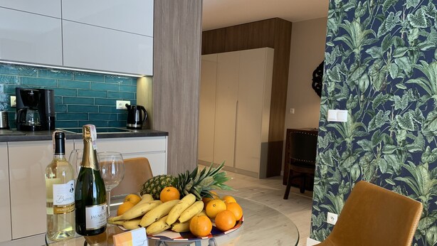 A modern kitchen with a coffee maker, a wine bottle, and a glass on the counter. There is a table with a glass, a bottle of wine, and a bowl of fruit. - ApartmentSun Parrot (Wroclaw)