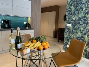 A modern kitchen with a coffee maker, a wine bottle, and a glass on the counter. There is a table with a glass, a bottle of wine, and a bowl of fruit. - ApartmentSun Parrot (Wroclaw)