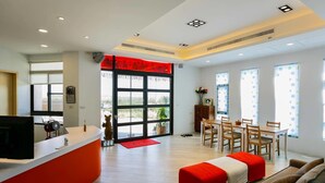 Een moderne hotellobby met een receptiebalie, een eetgedeelte met houten stoelen en een tafel, een rode accentmuur en grote ramen met gordijnen.