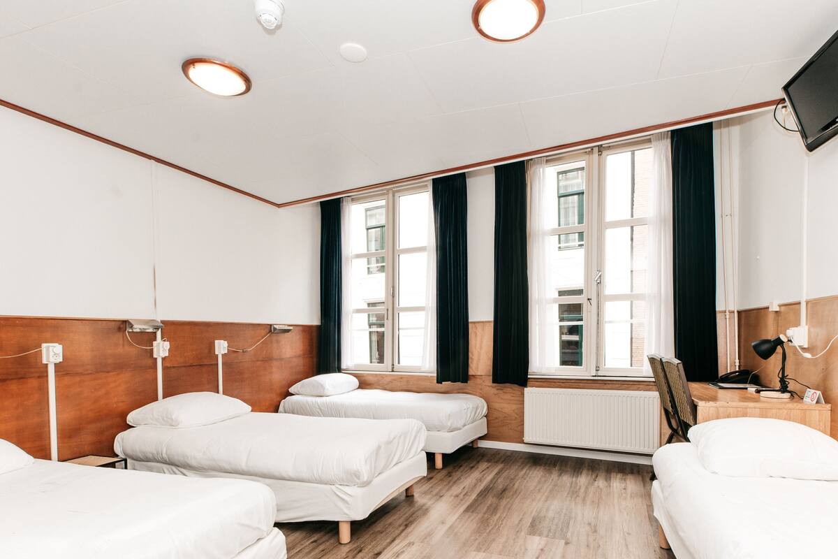 A hotel room with three single beds, wooden headboards, a large window with curtains, a radiator, a desk with a lamp, and a TV mounted on the wall.