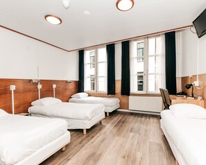 A hotel room with three single beds, wooden headboards, a large window with curtains, a radiator, a desk with a lamp, and a TV mounted on the wall. - Hotel van Gelder (Amsterdam)