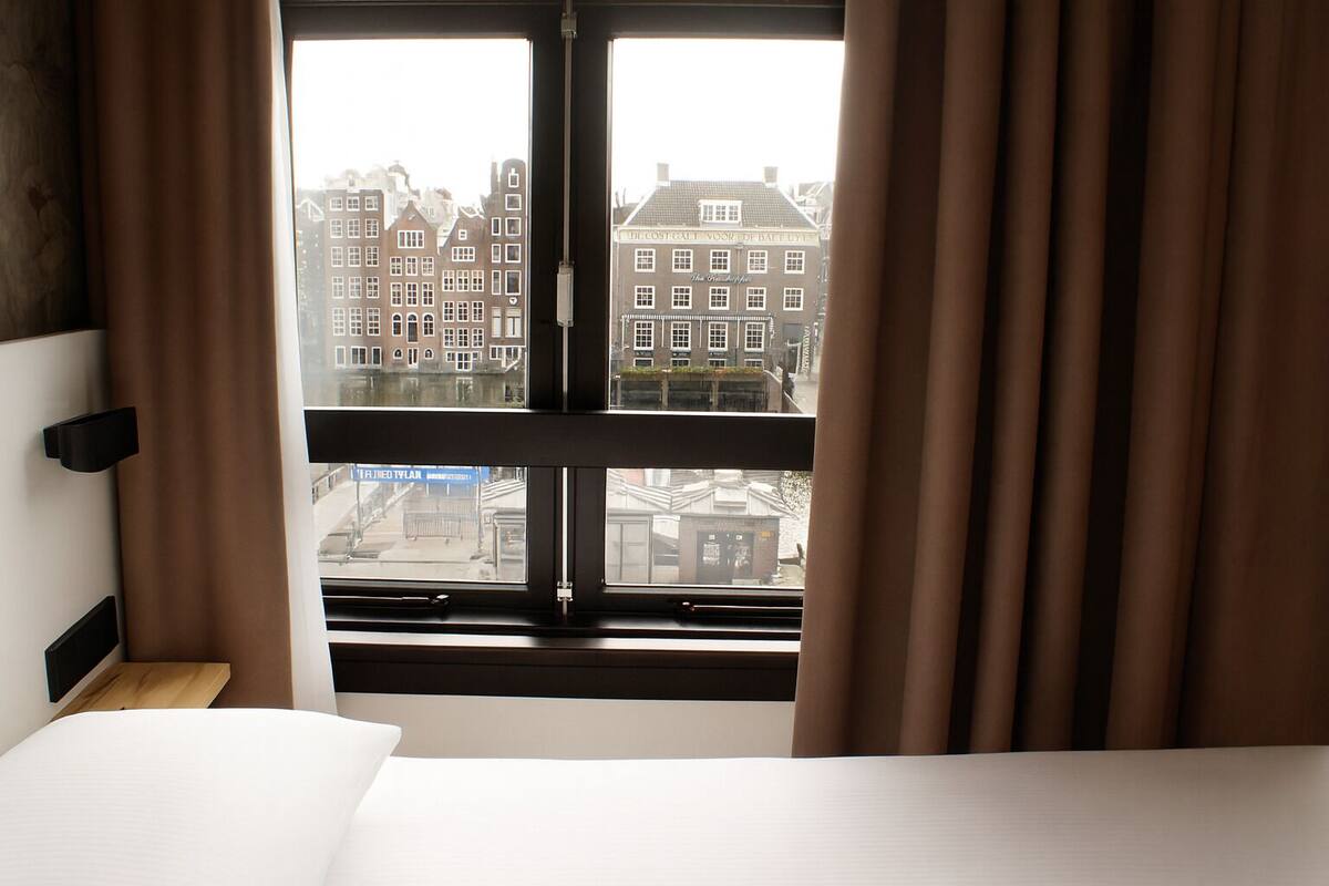 A hotel room with a bed, a window with curtains, and a view of buildings outside.