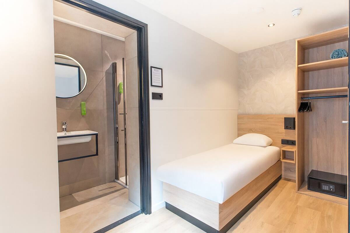 A compact hotel room with a shower, a small sink, a bed, and a wardrobe.