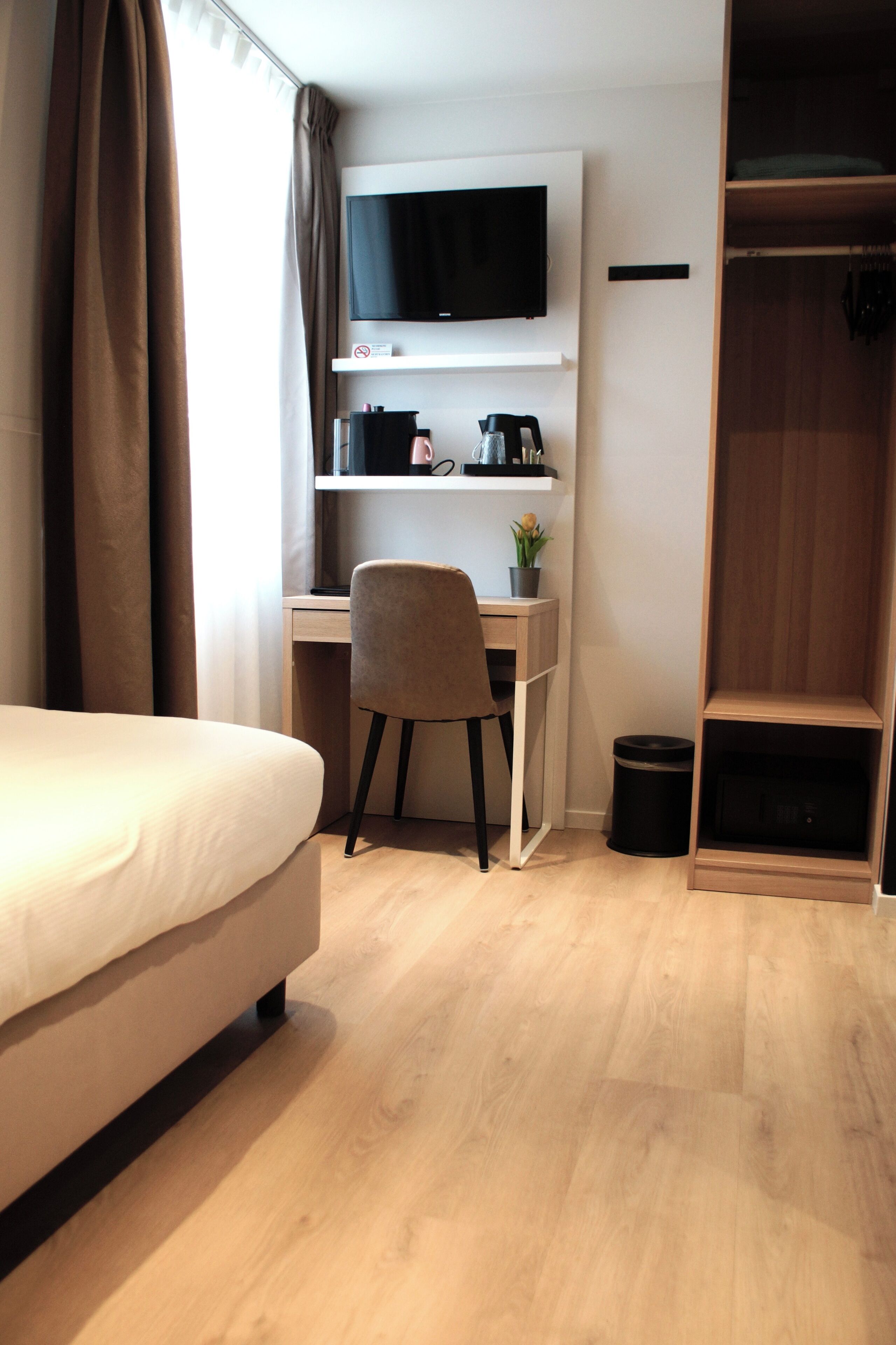 A hotel room with a bed, a desk with a chair, a television, a shelf with items, and a wardrobe.