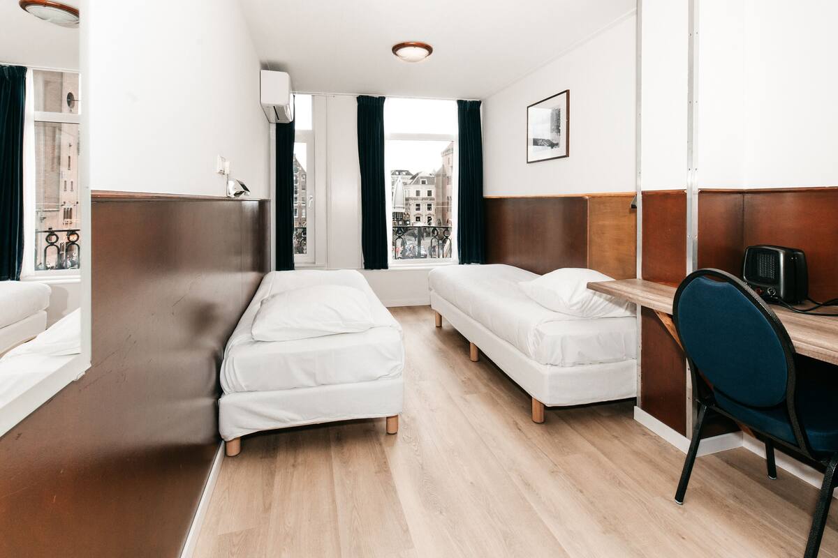 A hotel room with two single beds, a desk with a chair, a window with curtains, and a view of buildings outside.