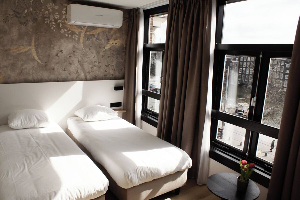 A hotel room with two beds, a window with curtains, and a view of buildings outside.