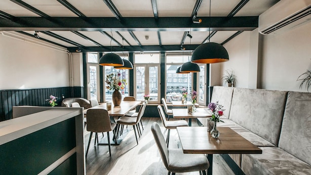 A modern restaurant interior with wooden tables and chairs, a bar area, and large windows offering a city view. - Hotel van Gelder (Amsterdam)