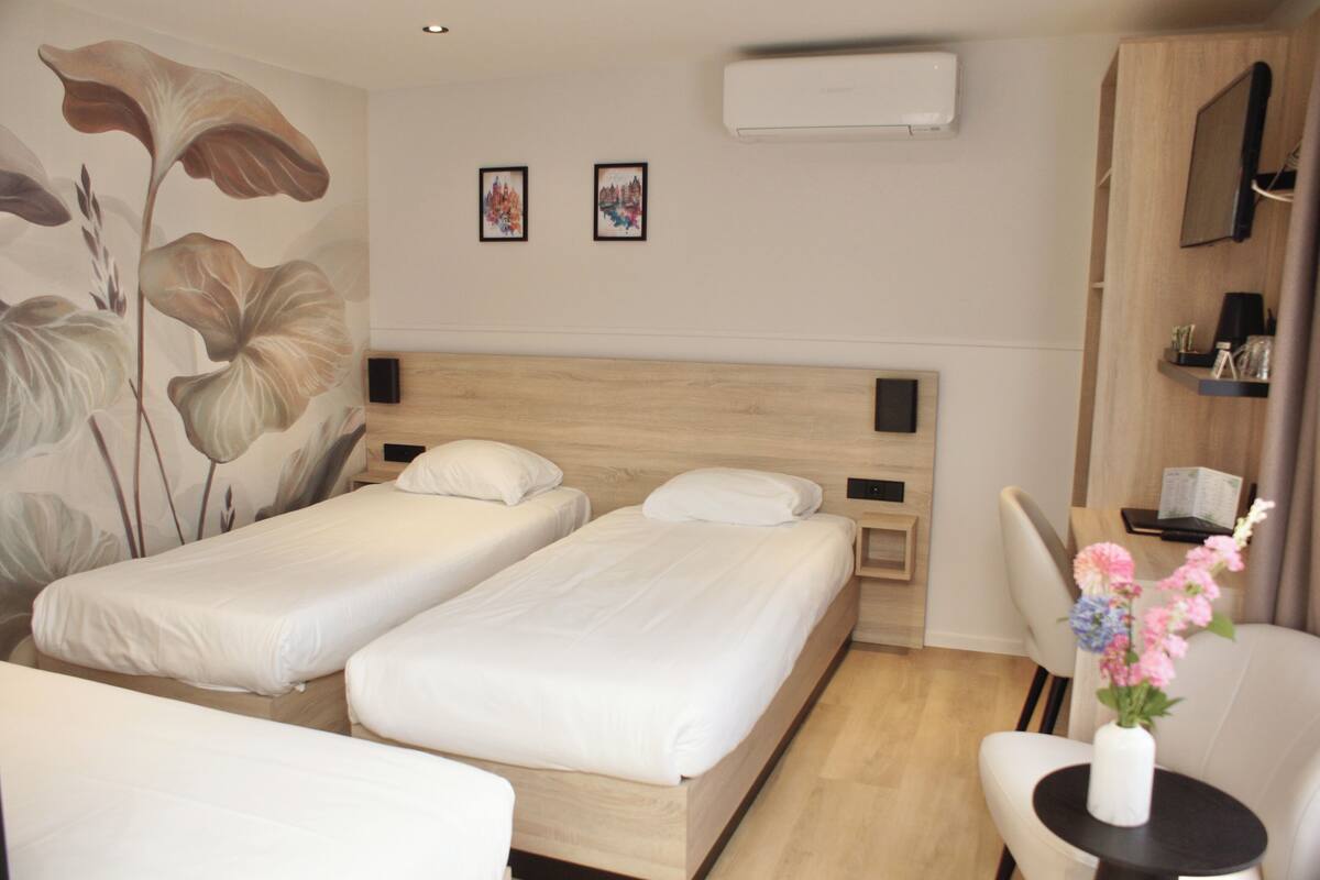 A hotel room with two single beds, a wooden headboard, a small table with a vase of flowers, a chair, and a wall-mounted TV.