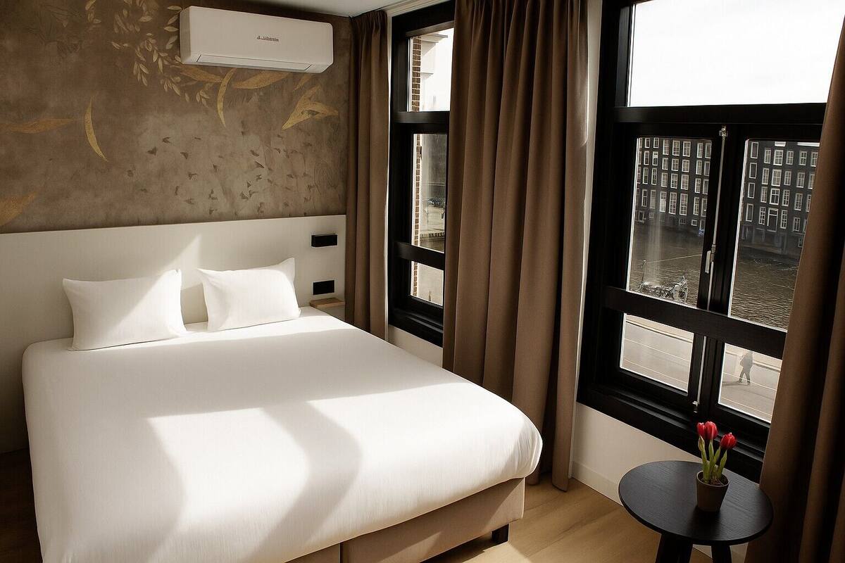 A hotel room with a large bed, a small table with a vase of flowers, and a view of buildings through large windows.
