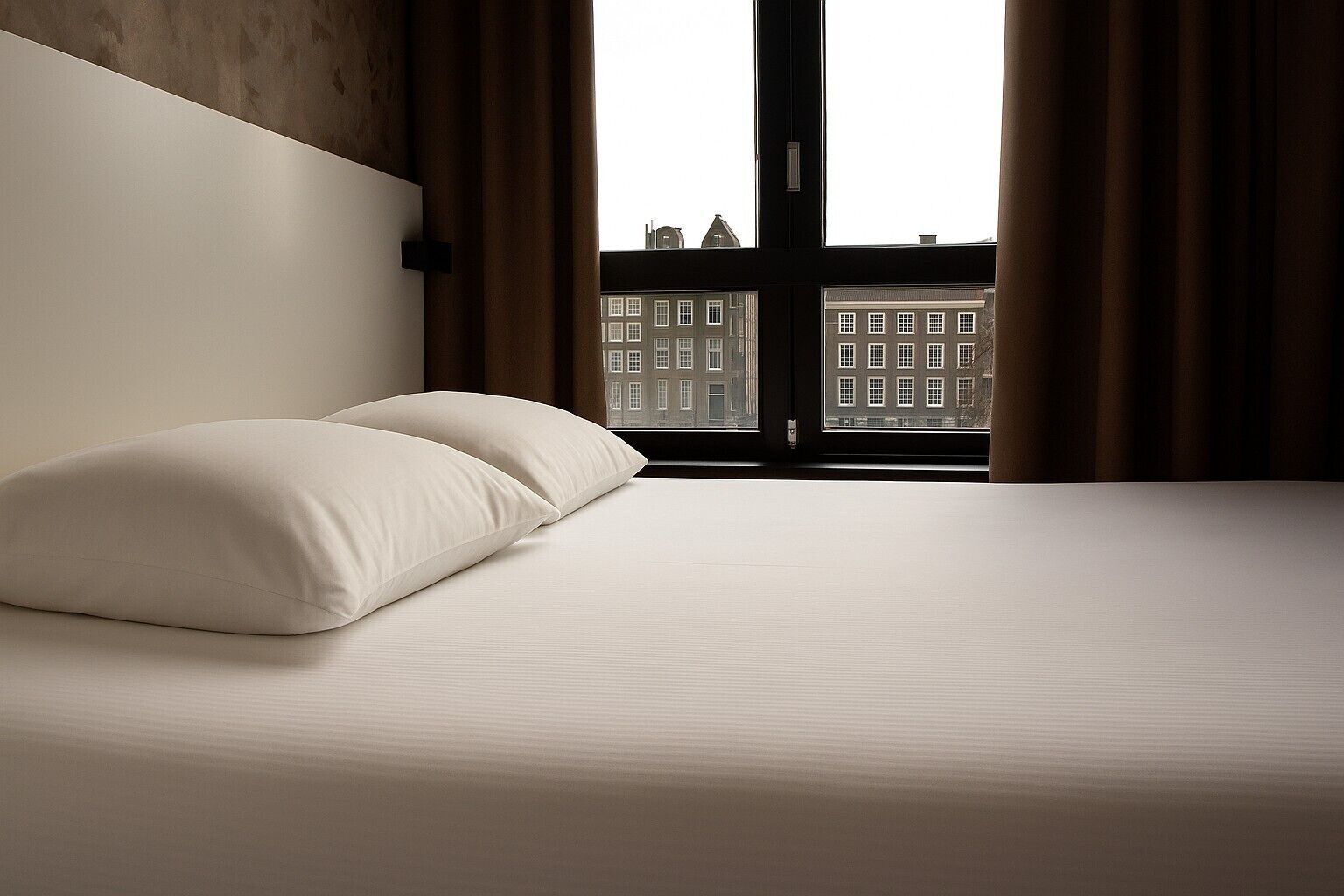 A hotel room with a large bed, two pillows, a window with curtains, and a view of buildings outside.