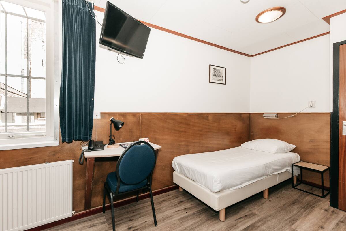 A room with a bed, a desk with a chair, a TV mounted on the wall, and a window with curtains.