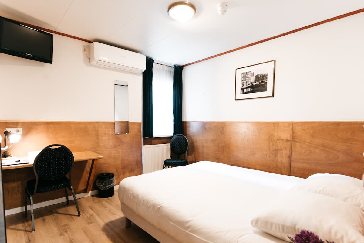 A hotel room with a wooden headboard, a bed with white linens, a desk with a chair, a mirror, a television, and a framed picture on the wall.