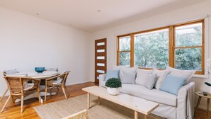 A bright living room with a white sofa, a round table, wicker chairs, and a large window with a view of trees. - Dovestones by the Sea - Inverloch (Inverloch)