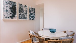 A dining area with a round table, wicker chairs, and three abstract paintings on the wall. - Dovestones by the Sea - Inverloch (Inverloch)
