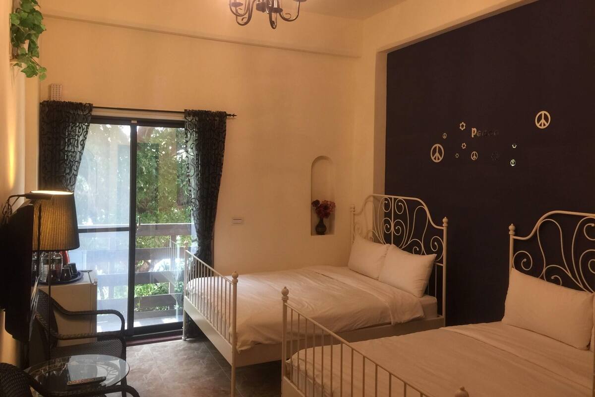 A room with two beds, a chandelier, a window with curtains, a small table, and a wall with peace symbols.